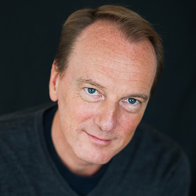 Marc Elsberg lächelt vor dunklem Hintergrund in die Kamera.