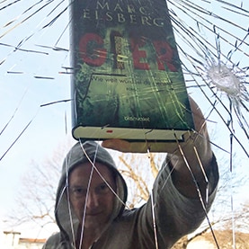 Marc Elsberg hält seinen Roman „Gier“, vor einer zerbrochenen Glasscheibe