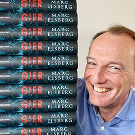 Marc Elsberg lächelt neben einm Stapel seines Romans „Gier“ in die Kamera.