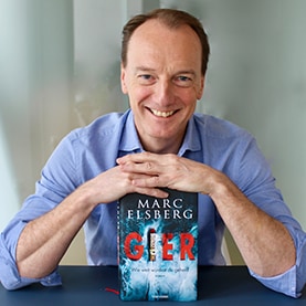 Marc Elsberg legt seine gefalteten Hände auf das Buch „Gier“, während er in die Kamera blickt.
