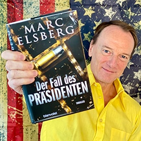 Marc Elsberg hält sein Buch „Der Fall des Präsidenten“ mit einer Hand vor der Amerikanischen Flagge.